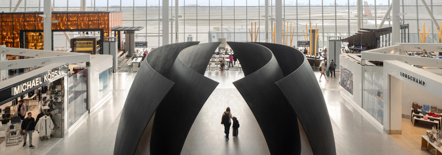 Interior view of a Toronto Pearson terminal with high ceilings and large windows allowing natural light to fill the space. In the center, there is a large, sculptural installation with curved, black structures. Surrounding the central installation are various high-end retail stores, including Michael Kors and Longchamp. Passengers are seen walking through the terminal, giving the space a lively atmosphere.
